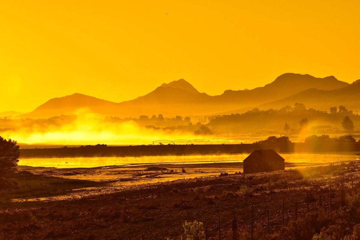 Gegenlichtaufnahme bei tiefstehender Sonne, durchgehend orange: Bergsilhouetten, Dunst über einer hellen Fläche, davor ein Schuppen, ein Weidezaun und eine Beregnungsanlage