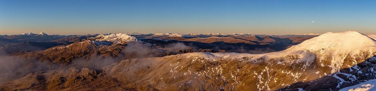 Sehr breites Panorama im Abendlicht: braune, teils verschneite Bergrücken mit Nebelfetzen bis zum Horizont, rechts ein Schneegipfel, darüber der Mond